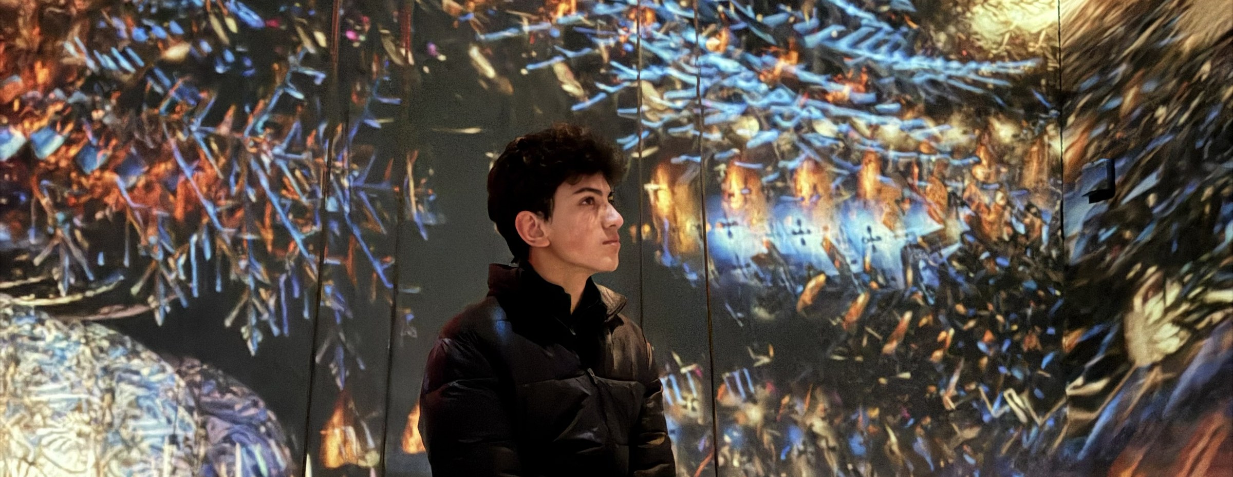 Aarav standing in profile inside an immersive light art installation, surrounded by warm and cool crystalline patterns